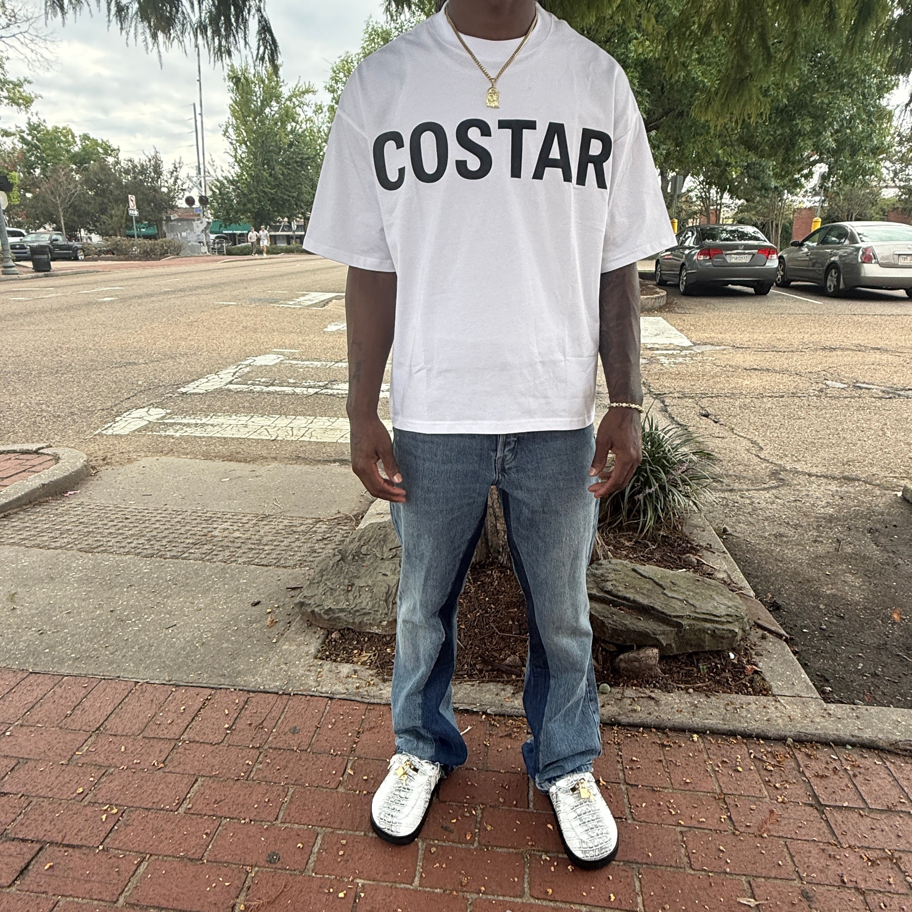 Person wearing a white t-shirt with 'COSTAR' printed on it, standing on a sidewalk.